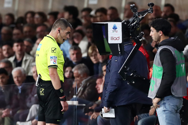 A football referee reviews a decision on the VAR monitor during a match, while a technician operates the screen beside him.