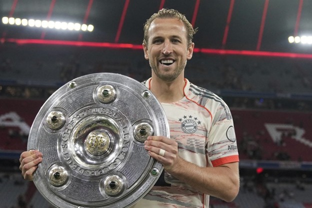 Harry Kane smiles while holding the Bundesliga championship shield on the pitch, celebrating his first major club trophy.