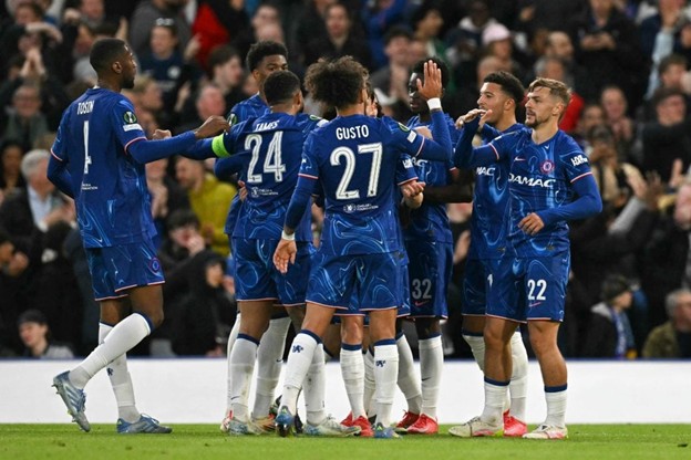 Chelsea players gather and celebrate after scoring a goal, with fans cheering in the background.