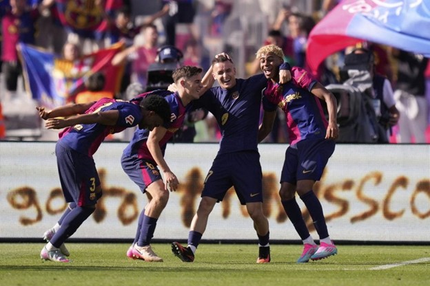 Barcelona players celebrate a goal on the pitch with joyful expressions and playful poses, surrounded by waving flags and cheering fans in the background.