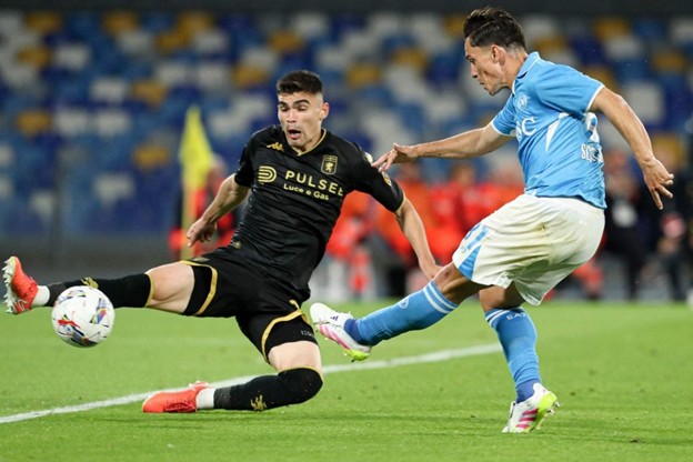 A Napoli player takes a powerful shot as a Lecce defender slides in to block during an intense moment of the match, both wearing their respective team kits.