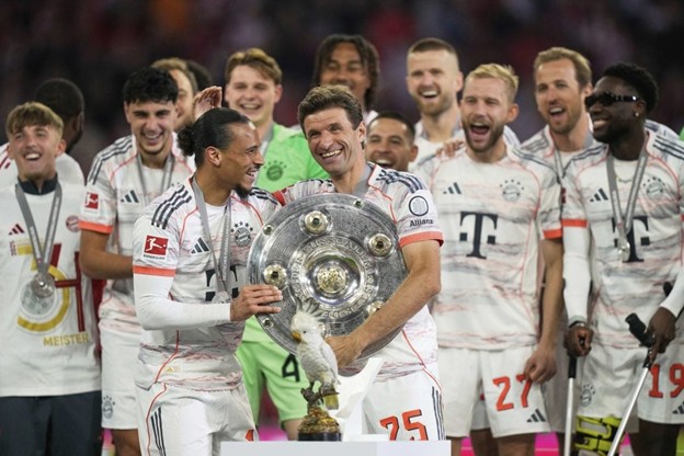 Bayern players celebrate with joy as Thomas Müller and Leroy Sané hold the Bundesliga trophy together, surrounded by teammates, with a golden eagle trophy in front of them.