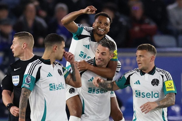 Newcastle United players celebrate together after scoring a goal during a match, with the team wearing their white away kit. One player jumps onto a teammate's back in joy.
