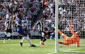 A Premier League match moment where a Newcastle United player scores past a diving goalkeeper, while a Chelsea defender watches helplessly, with fans in the background.